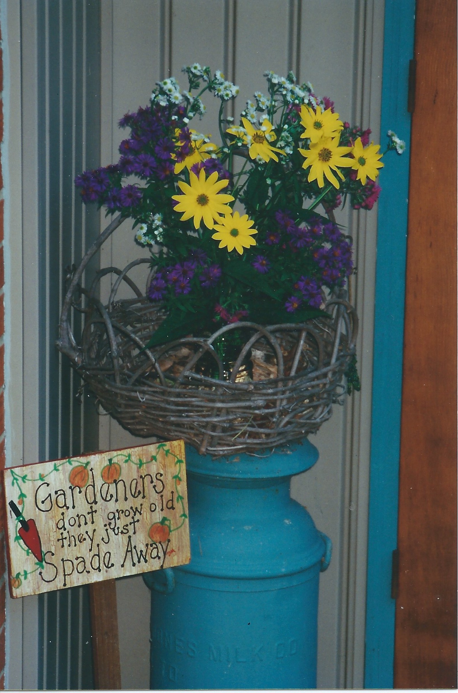 Blue milk can with basket of flowers and sign that says Gardeners don't grow old - they just Spade Away.