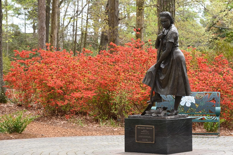 A bronze statue titled 'Breaking Ground' honoring workers in the Norfolk Botanical Garden, set against a backdrop of vibrant red azalea flowers.