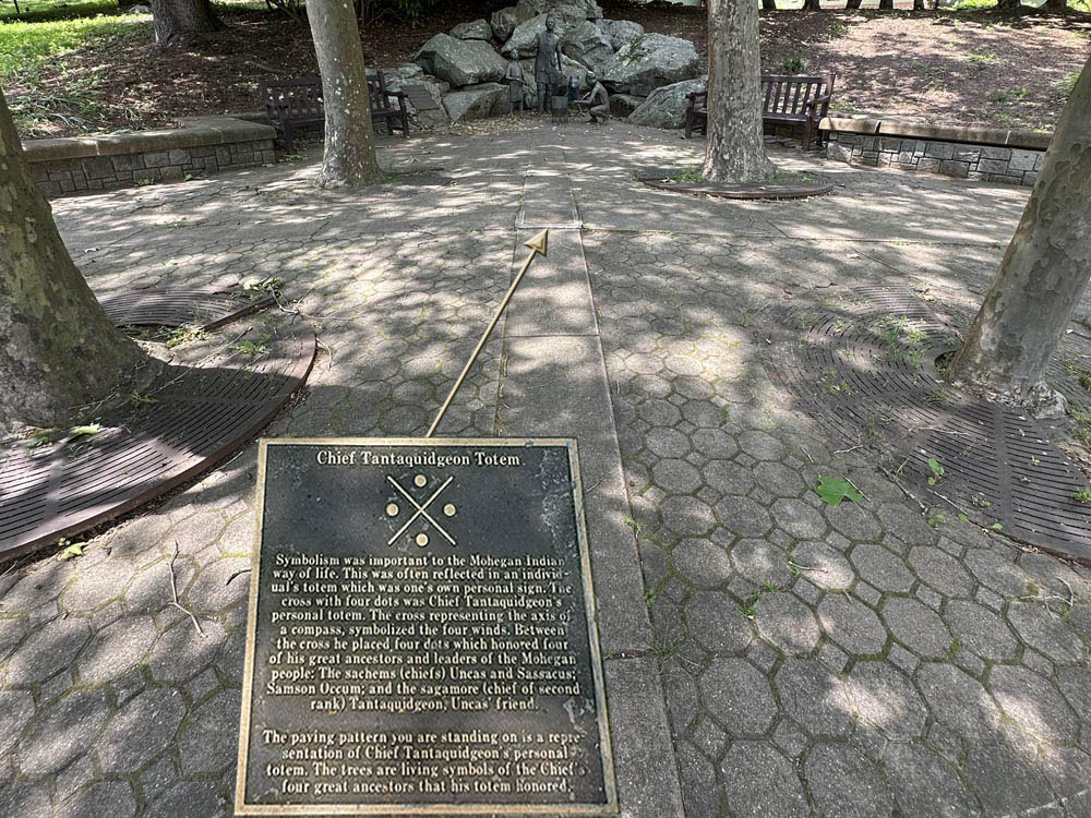 A plaque at the center of a paved representation of Chief Tantaquidgeon’s Totem<br/> explains that the rectangular pavers depict the cross and the four trees, the dots.<br/>At the far end is the 1994 Mohegan Heritage Sculpture..