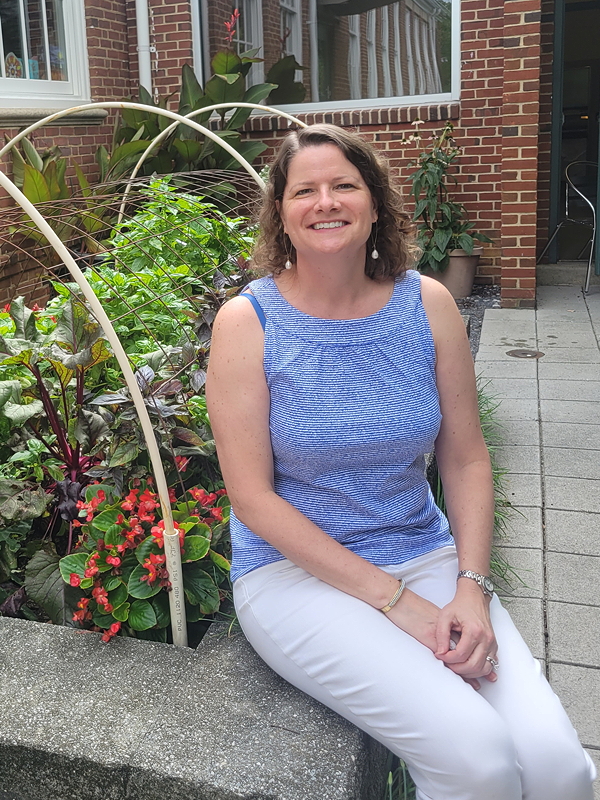 A woman sitting on a concrete ledge in a garden, wearing a blue sleeveless top and white pants, smiling at the camera. Behind her are various plants and a brick wall.