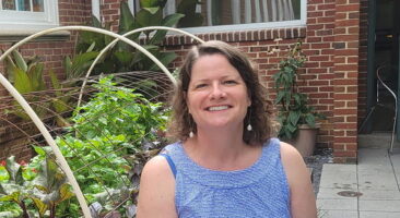 A woman sitting on a concrete ledge in a garden, wearing a blue sleeveless top and white pants, smiling at the camera. Behind her are various plants and a brick wall.