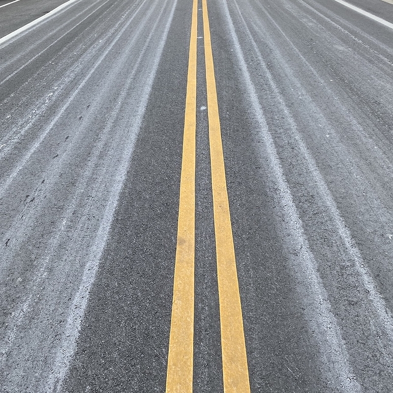 View of a road from above with two parallel yellow lines showing a textured grey surface with white salt treatment lines.