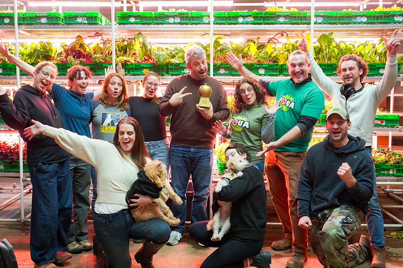 A group of people celebrating in an indoor farm, holding a golden trophy, with plants in the background.