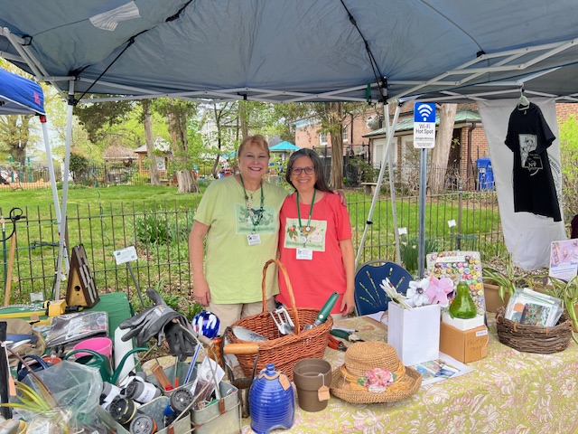 Two women standing together at a craft fair booth under a canopy, smiling at the camera, surrounded by various items for sale, including gardening tools and handmade crafts.