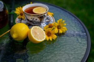 A cup of tea with a slice of lemon is placed on a decorative saucer, surrounded by fresh lemons and vibrant yellow flowers on a glass table.