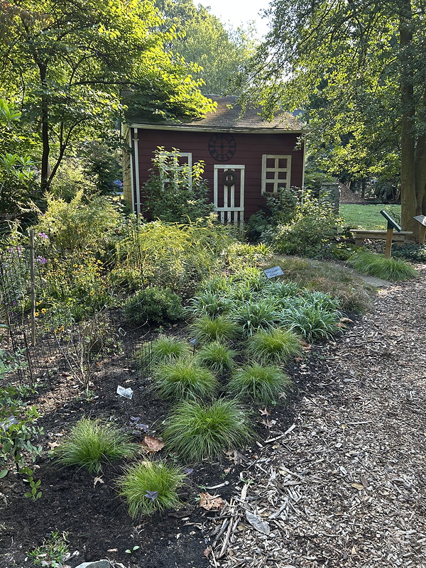 A small red house surrounded by lush green plants and flowers in a garden setting.