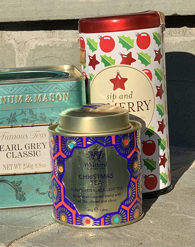 A collection of decorative tea tins including a blue tin labeled 'Earl Grey Classic', a gold tin for 'Christmas Tea', and a festive red and green patterned tin labeled 'sip and merry'.