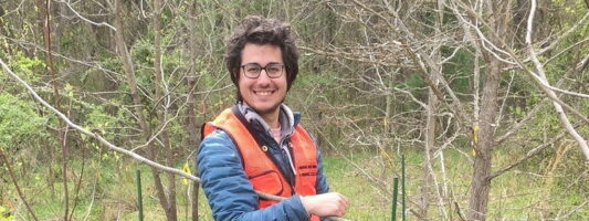 A young man smiles while standing outdoors in a forested area, wearing an orange safety vest and glasses. He is positioned between two black insect trapping tents surrounded by greenery and bare trees.