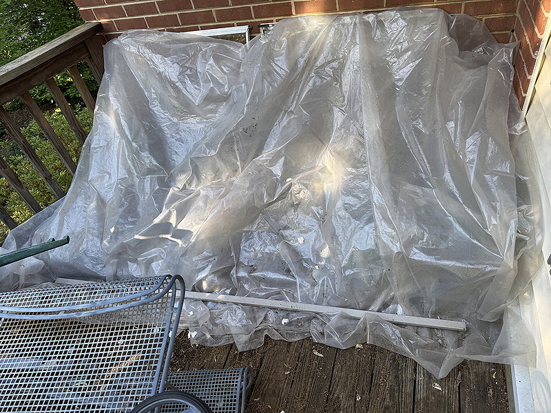 A covered outdoor patio area with a clear plastic tarp draped over a wooden surface, accompanied by metal furniture components.